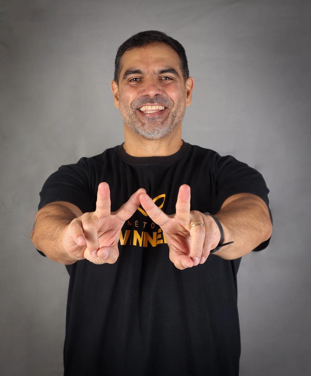 Homem branco de cabelo preto, com blusa preta fazendo sinal de paz com as duas mãos esticadas.