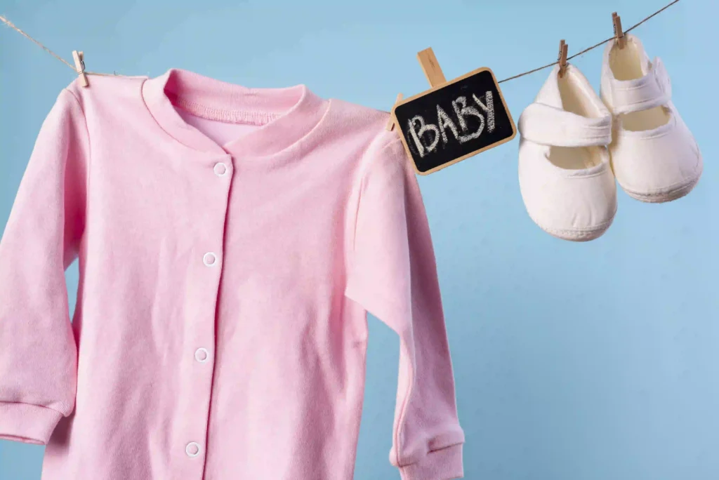 Imagem com fundo azul claro, simulando uma varal com casaco infantil rosa, placa escrito "baby" e sapatinho branco" pendurados.