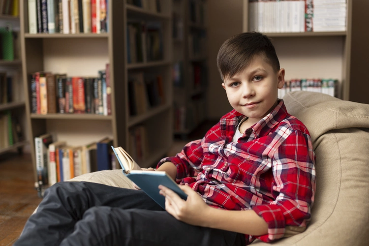 Imagem de um menino branco com cabelo castanho liso e franja lateral, vestindo uma blusa quadriculada vermelha e branca, sentado em um pufe bege em uma biblioteca ao fundo.