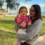 Mulher branca de óculos com sua filha pequena no colo em um local aberto e gramado.