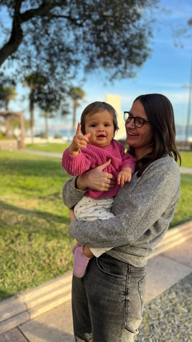 Mulher branca de óculos com sua filha pequena no colo em um local aberto e gramado.