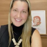 Mulher loira de cabelos longos e blusa preta com um cordão de pingente grande e com o rosto sorrindo, com uma parede amadeirada ao fundo e um quadro pequeno branco