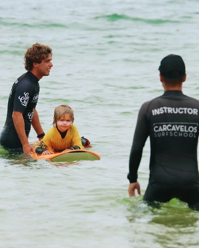 Criança pequena loira com blusa amarela sob uma prancha com o auxilio de um instrutor de uniforme preto