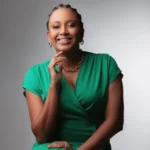 Mulher negra sorrindo sentada usando uma roupa verde e posando com a mão no queixo com a qual tem um anel verde