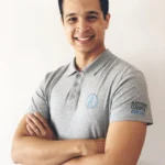 fundo branco. fotografia de um homem a sorrir, polo de fisioterapeuta cinzento. Pele morena, cabelos pretos curto, olhos rasgados. Encontra-se de braços cruzados com a luz natural à refletir o corpo.