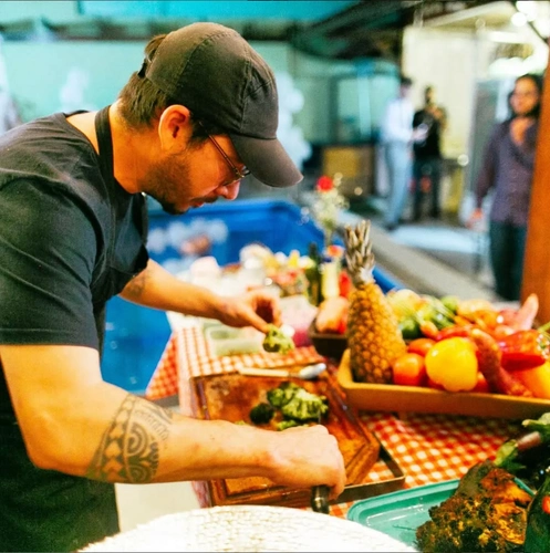 homem branco de boné, blusa e avental na cor grafite, usando óculos de armação vermelha e picando alimentos sobre uma mesa com cestos de frutas. Imagem de 3 pessoas ao fundo desfocada.