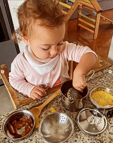 Menina branca de pequena de roupa rosa e babador branco, sentada em uma cadeira e com as mãos apoiadas em balcão enquanto prepara comida em potinhos de aço inox. 