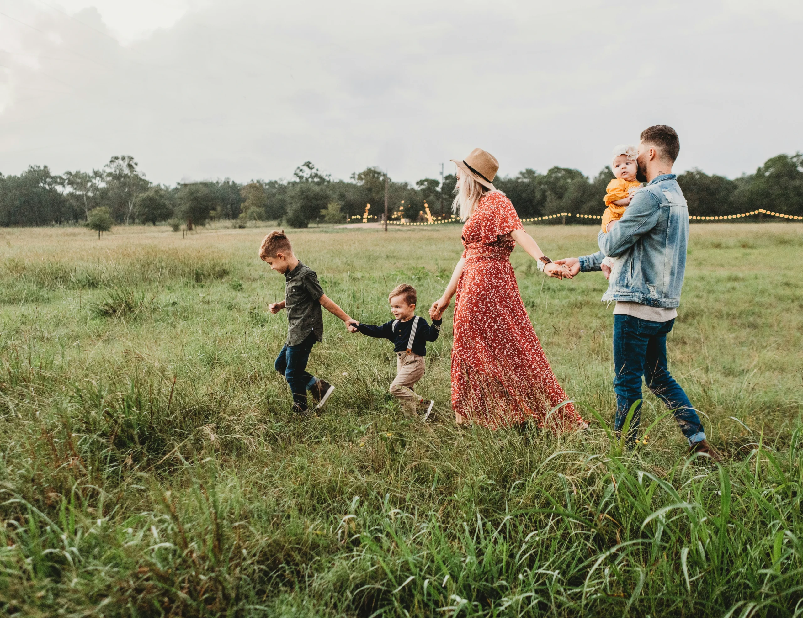 família de casal com três filhos de mãos dadas no campo. o céu está azul e claro. Menino mais velho em primeiro lugar de cabelo castanho claro e roupa azul escura. logo atrás vem o irmão do meio com as mesmas caracteristicas mas mais novo. Este dá a mão à mâe, loira de cabelos compridos com um vestido florido vermelho e chapeu castanho. Esta dá a mão para o esposo em último lugar de cabelo castanho escuro, jaqueta jeans e calças jeans segurando o bebe mais novo no colo.
