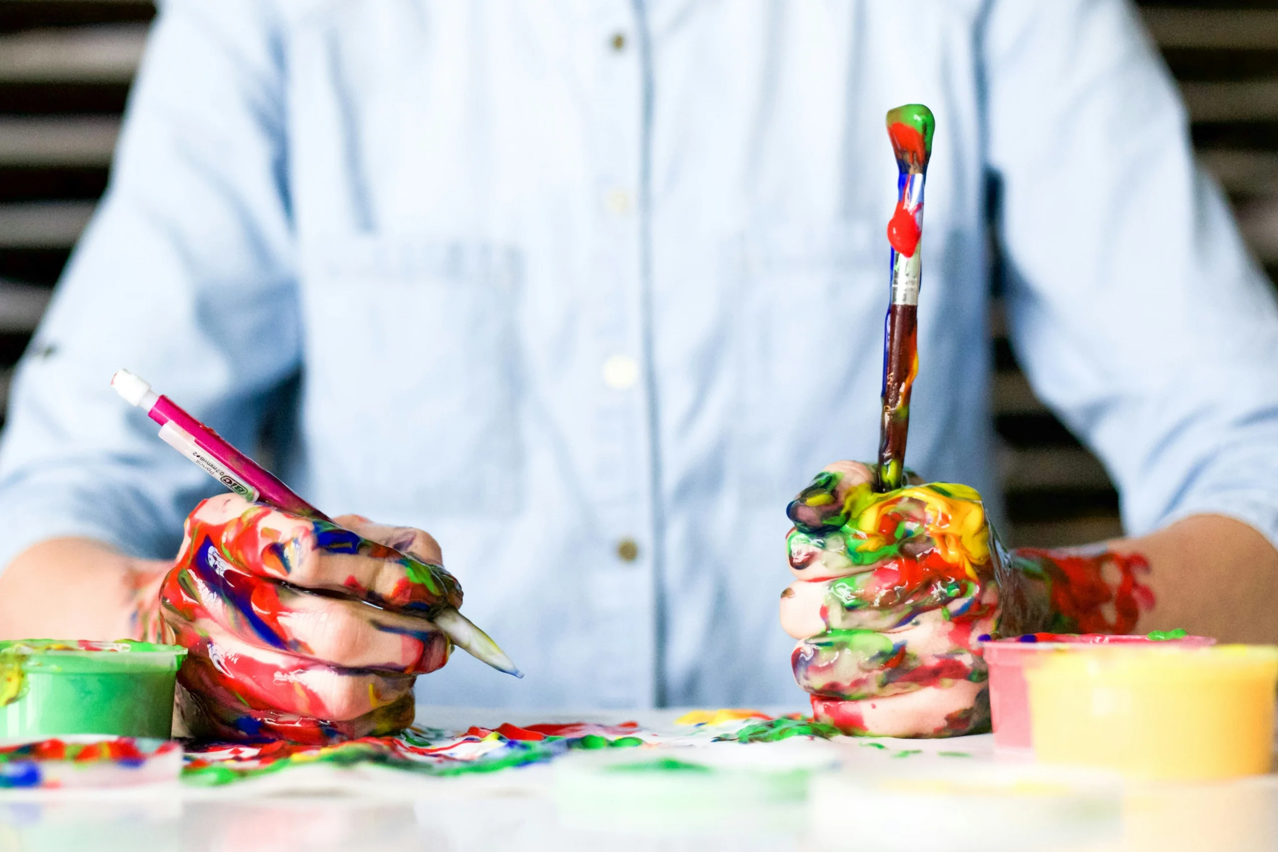 Menino com camisa azul clara e com as mãos pousadas sobre uma mesa. As mãos estão segurando utensílios de arte e estão sujas de tinta. A mesa está bem suja de tinta também. Não se vê o rosto.