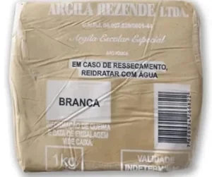 Pacote de 1 kg de argila branca escolar da marca "Argila Rezende Ltda." com instruções em português. O rótulo informa: "Em caso de ressecamento, reidratar com água", e destaca a palavra "BRANCA" indicando a cor da argila. Embalagem de plástico transparente mostrando a textura da argila.
