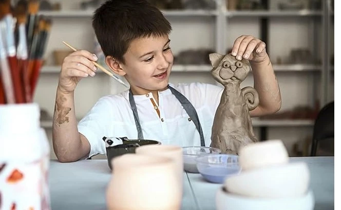 Menino sorridente sentado em uma mesa de artesanato, moldando uma escultura de gato com argila. Ele segura um pincel em uma mão e a escultura na outra. Ao fundo, há prateleiras com materiais de arte e cerâmica. A criança está se divertindo durante uma atividade artística.