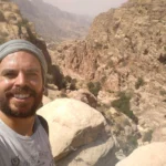 Rafael Polónia sorrindo durante uma trilha nas montanhas áridas da Jordânia, aproveitando um momento de conexão com a natureza, simbolizando o equilíbrio entre pai e tempo.