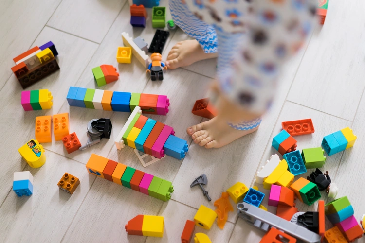 Pés descalços de uma criança e de um adulto sobre piso claro, cercados por blocos de montar multicoloridos e pequenas figuras espalhadas, sugerindo momento de brincadeira colaborativa no chão