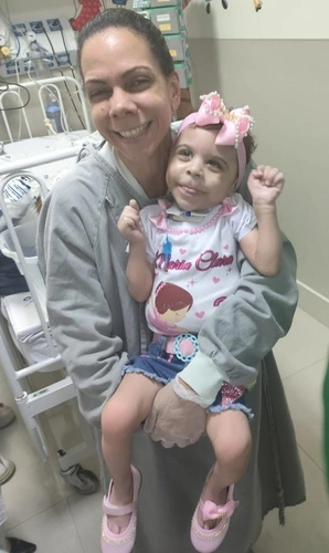 Mulher de pele clara, cabelos escuros presos e vestindo jaleco cinza, sorri enquanto segura uma menina pequena com síndrome congênita, pele clara, usando faixa rosa na cabeça, camiseta branca estampada com desenho infantil e shorts jeans, dentro de ambiente hospitalar com equipamentos ao fundo.