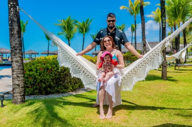 Família sorrindo em rede de descanso, com jardim, coqueiros e céu azul ao fundo.