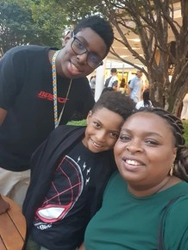 Mulher negra de pele escura e cabelos trançados sorrindo, sentada ao ar livre ao lado de dois meninos negros, um adolescente de óculos e um mais novo, todos sorrindo, em ambiente de shopping com árvores ao fundo.