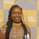 Mulher negra de pele escura, com cabelo trançado e colar dourado, sorrindo em frente a um painel de fundo amarelo e cinza com a frase “Mães Negras” repetida várias vezes, usando vestido estampado colorido.