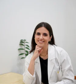 Mulher jovem de cabelo castanho escuro e liso, sentada junto a uma mesa de madeira clara, a sorrir para a câmara. Usa bata branca de profissional de saúde, uma blusa preta e calças claras. Tem brincos dourados, um colar dourado com uma letra "P" e está com a mão apoiada no queixo. Ao fundo, vê-se uma parede branca e uma planta decorativa