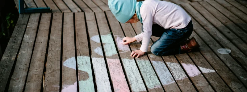 menina desenhando corações com giz de cera colorido no chão de madeira exterior. a menina está debruçada no chão a desenhar com uma camisa rosa e touca azul clara.