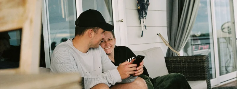 Casal em segurança sentado na varanda de casa inteligente felizes a olharem para o smartphone. ELa está com uma manta verde escura e a casa é branca de madeira.