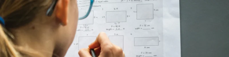 Uma criança com cabelos presos e óculos está concentrada em resolver um exercício de matemática em uma folha de papel. A imagem mostra a visão de trás, com a criança segurando um lápis e escrevendo as respostas. O exercício envolve cálculo de perímetro e medidas desconhecidas de retângulos. O ambiente tem uma iluminação suave, e a mesa cinza contrasta com o papel branco da atividade.