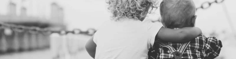 Foto em preto e branco. Cidade no fundo num cais. Duas crianças sentadas no chão de costas abraçadas. Os dois com menos de dois anos, o da esquerda, mais velho, loiro com cabelo encarracolado e camisa branca. O mais novo de cabelo claro e roupa escura