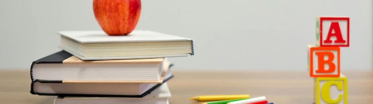 Mesa de um professor com livros, uma maça, alguns lápis e cubos com letras.