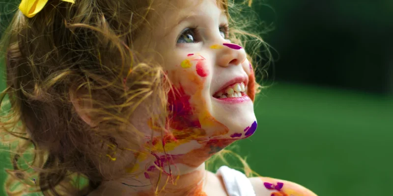 Criança sorridente com cabelos cacheados e laço amarelo, com o rosto, braços e roupa cobertos por tinta colorida, em um ambiente ao ar livre.